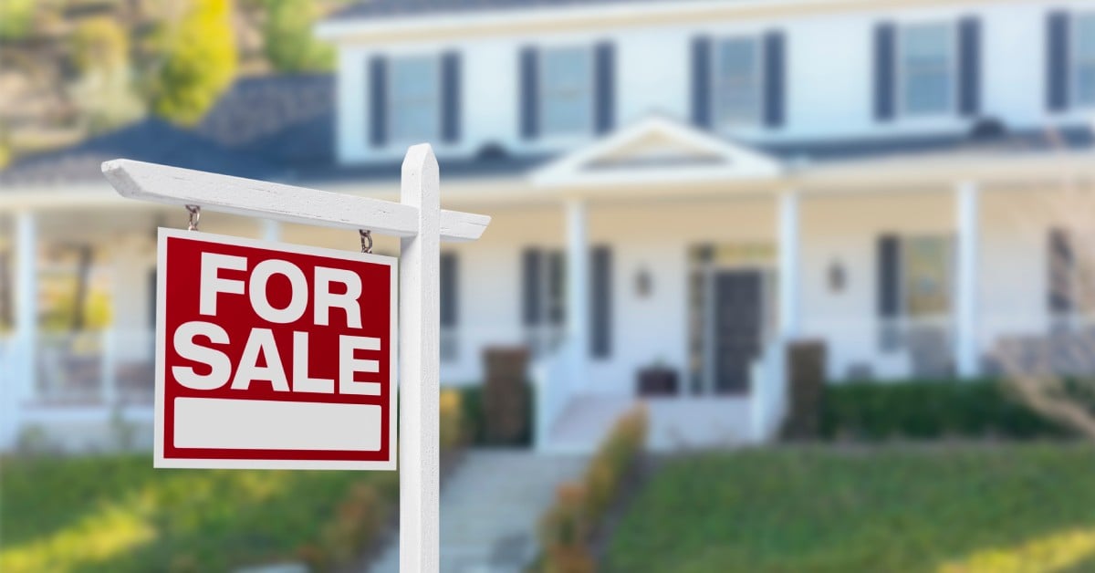 A red and white FOR SALE sign hangs on a white post in front of a two-story house with white siding and dark shutters.