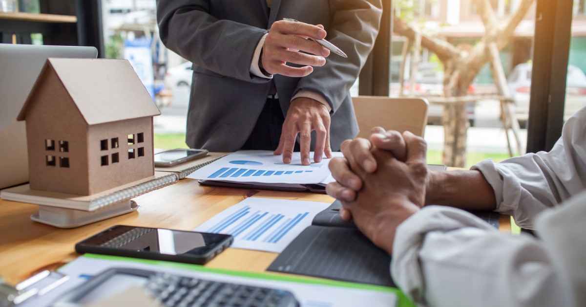 One person standing and pointing at a document while another sits beside a model house and office supplies on a table.
