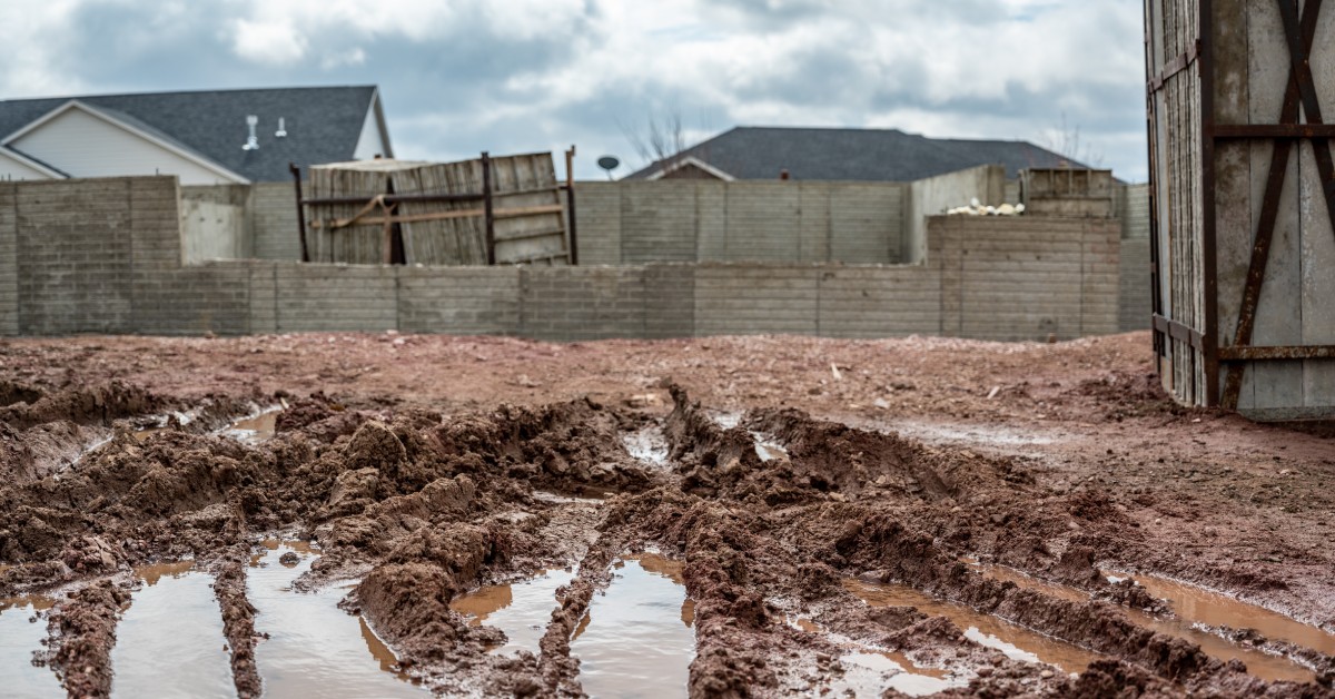 A muddy construction lot showing deep tire tracks and puddles, with unfinished block walls and houses behind.