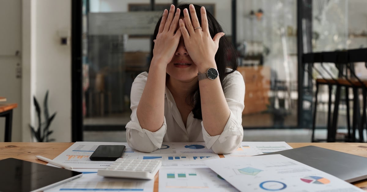 A woman sitting at a desk covered with charts, papers, a calculator, and devices while covering face with both hands.