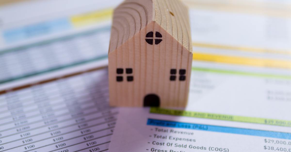 A mini wooden house model sitting on financial spreadsheets with revenue, expenses, and profit details visible.