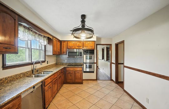 kitchen, double wall oven, cooktop, built in microwave, dishwasher, granite countertops, cabinets, tile flooring, sink, window above sink, pot rack light, wood trim