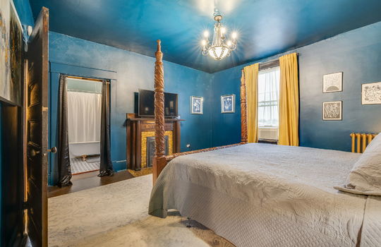 primary bedroom, fireplace, tile surround on fireplace, wood mantle, hardwood flooring, window, chandelier, doorway to ensuite bathroom, radiator