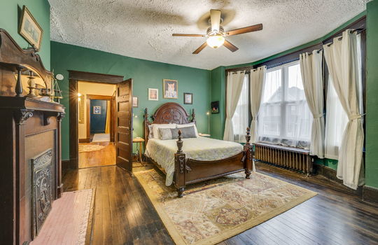 bedroom, fireplace, hardwood flooring, wood trim, bay window, radiator, fireplace, wood mantle, wood door, ceiling fan