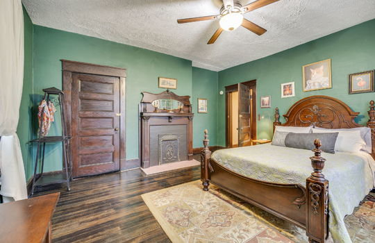 bedroom, fireplace, hardwood flooring, wood trim, bay window, radiator, fireplace, wood mantle, wood door, ceiling fan