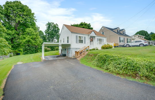 left side view of home, one story, cottage, carport, driveway, stairs front porch, vinyl siding, shingle roof, front door, fencing, trees, front yard