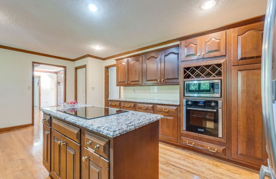 kitchen, kitchen island, cooktop, cabinetry, granite countertops, built-in microwave, built-in oven, tile backsplash, wine rack, hardwood flooring, pantry