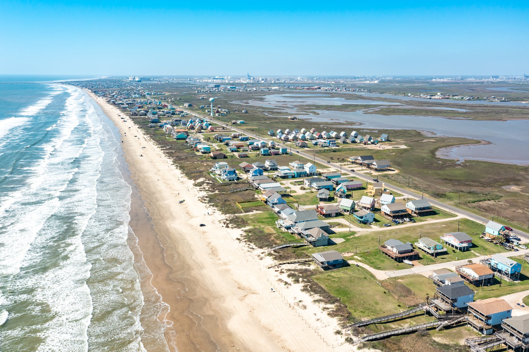 Drone view of Surfside Beach, Texas, showing beachfront homes lined along the coast with the Gulf of Mexico waves on one side and marshland on the other.