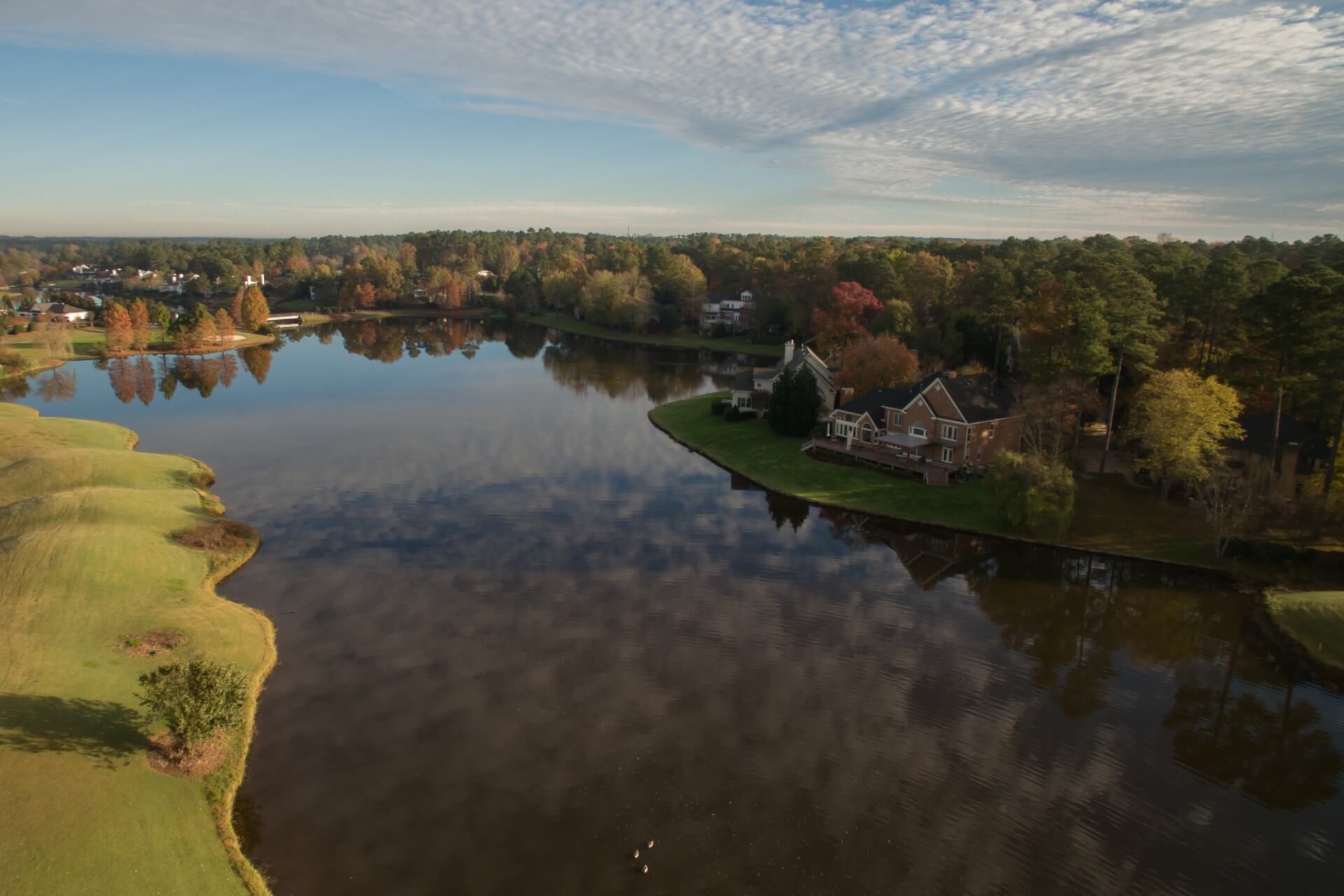 Aerial view of a lake bordered by a golf course and a brick home with fall trees in Cary, NC