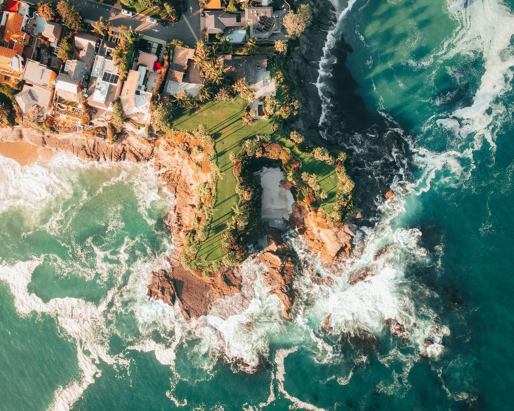 Drone view of homes on a rocky coastal hill above turquoise surf