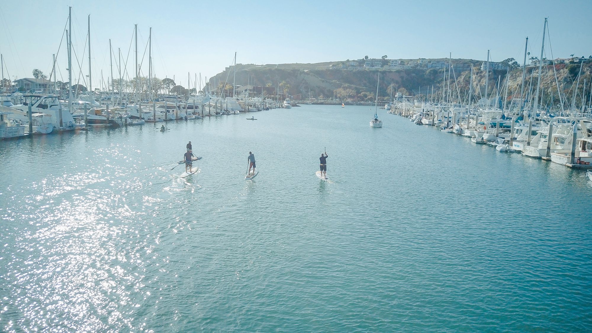 Paddleboarders crossing a sunny marina flanked by moored yachts