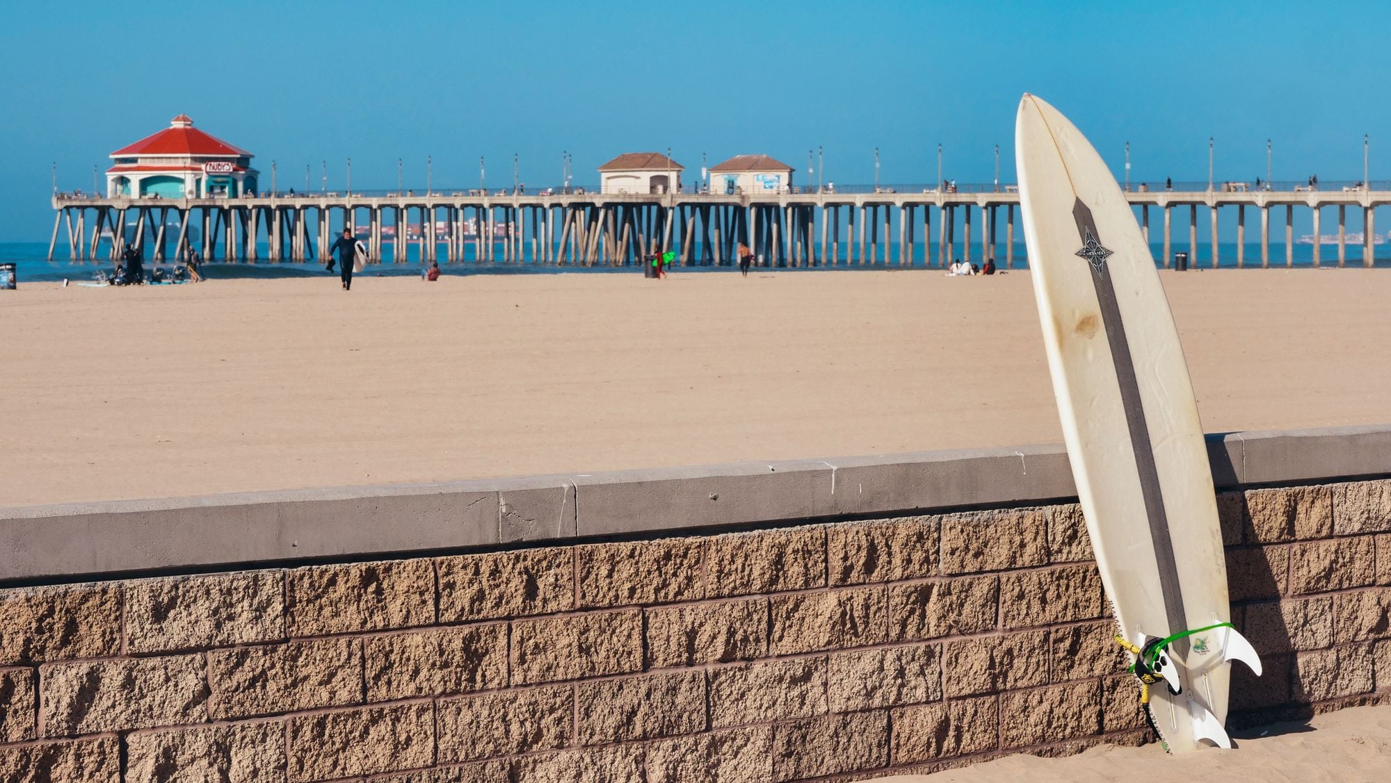 A surfboard leaning on a low wall with a beach pier behind it