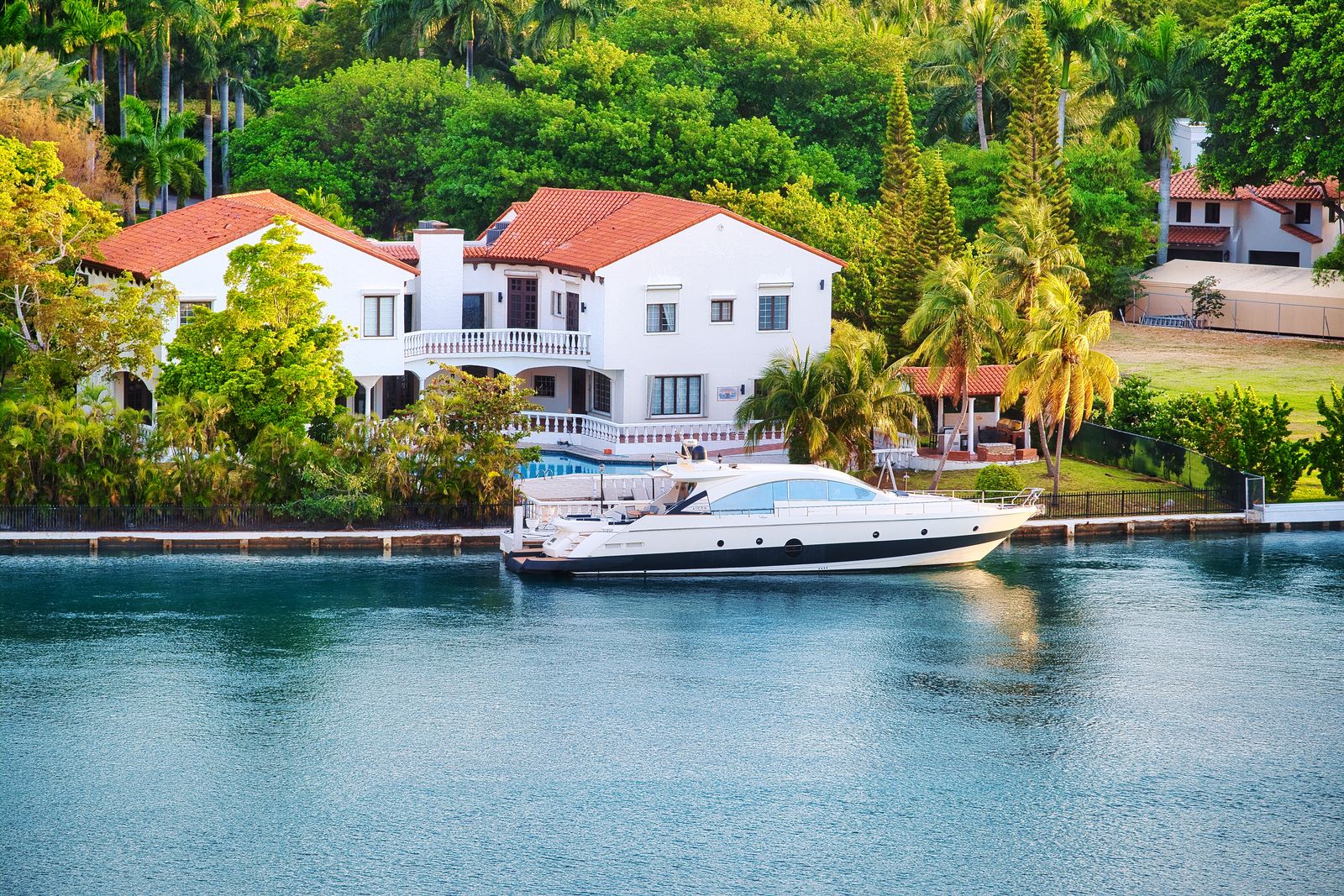 A white waterfront villa in Miami with a motor yacht moored at its private dock, palm trees lining the seawall