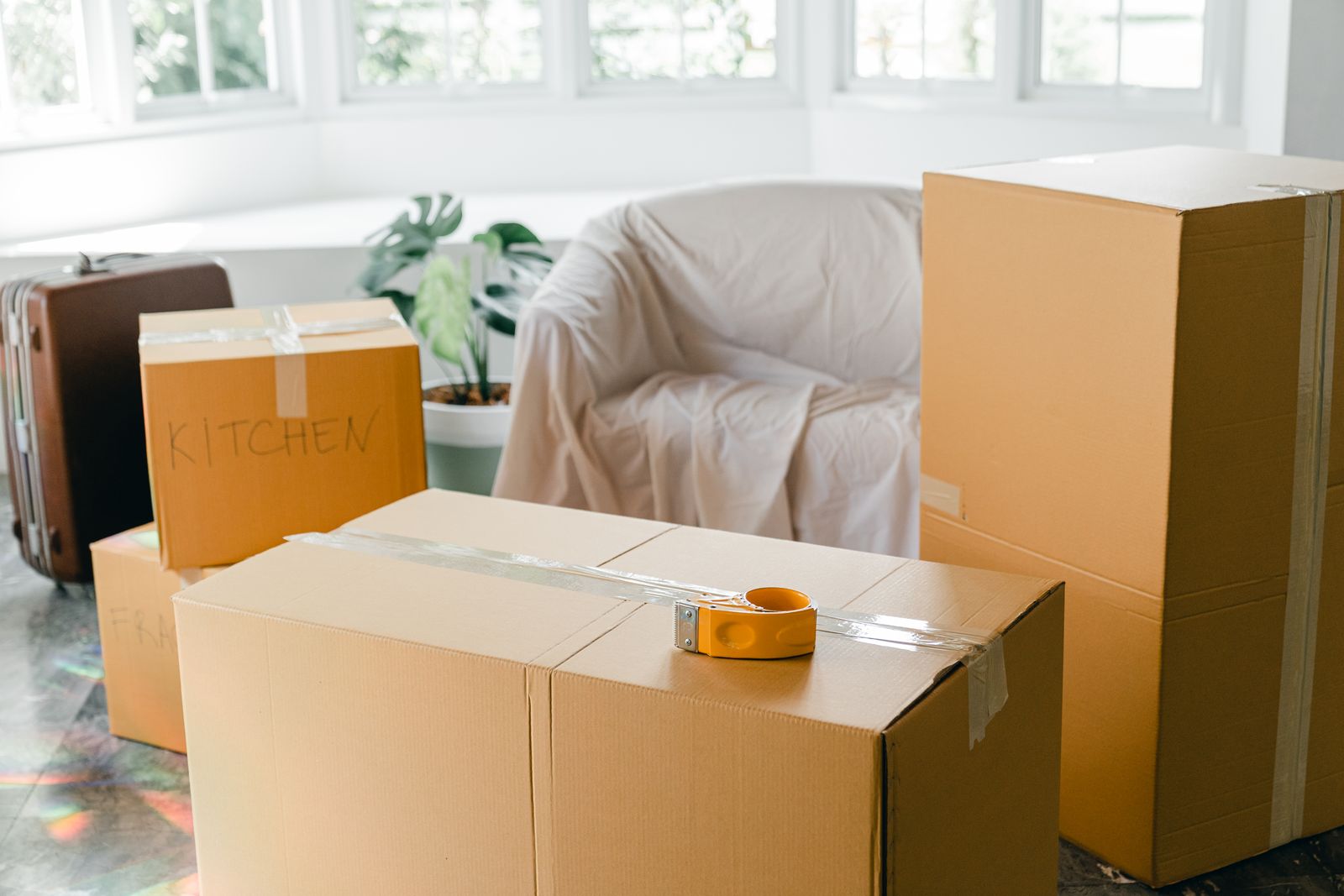 Cardboard moving boxes labeled Kitchen stacked on the floor of a sunlit living room before a relocation
