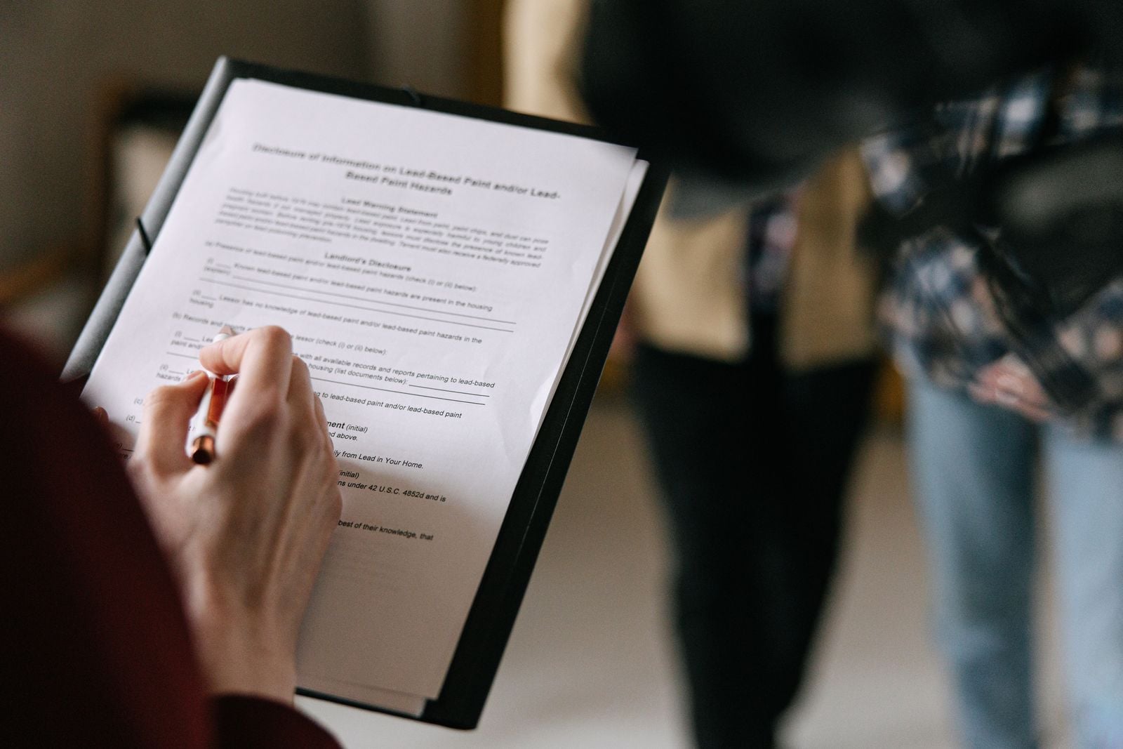 Woman filling out a property disclosure form on a clipboard while two people stand talking behind her
