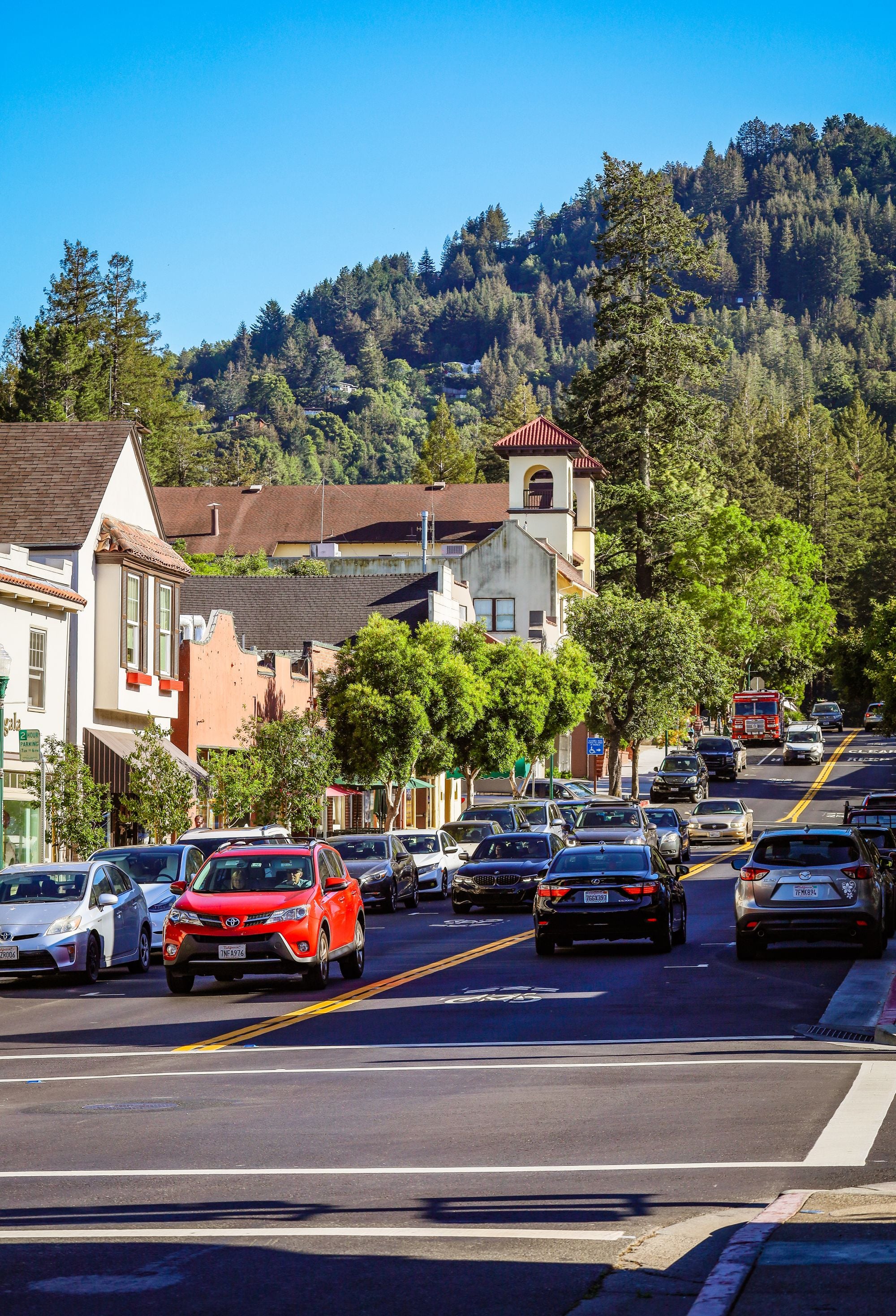 A small town street lined with shops and parked cars below low hills
