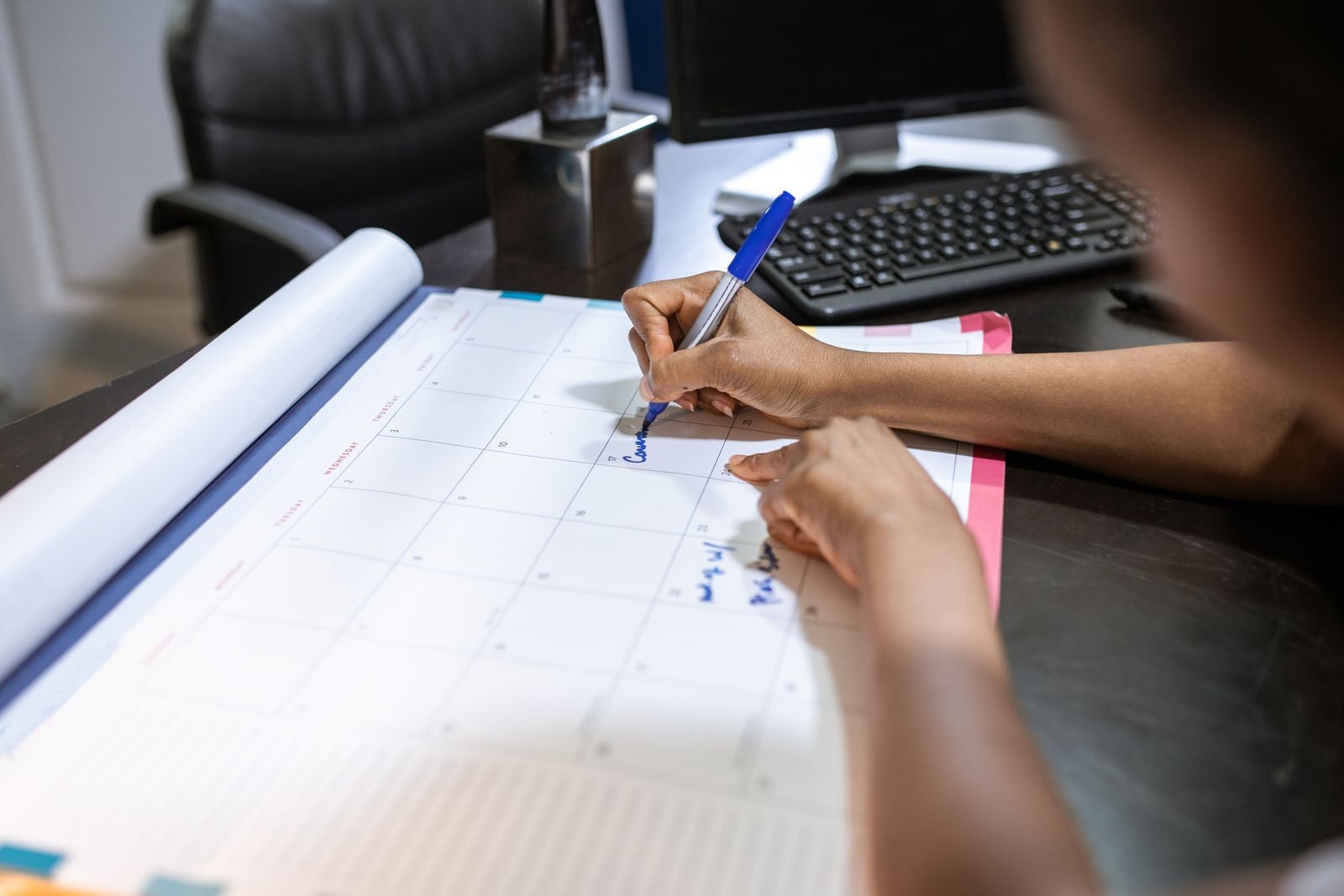 A person writing on a large paper desk calendar with a blue pen, with a keyboard and a rolled document beside it