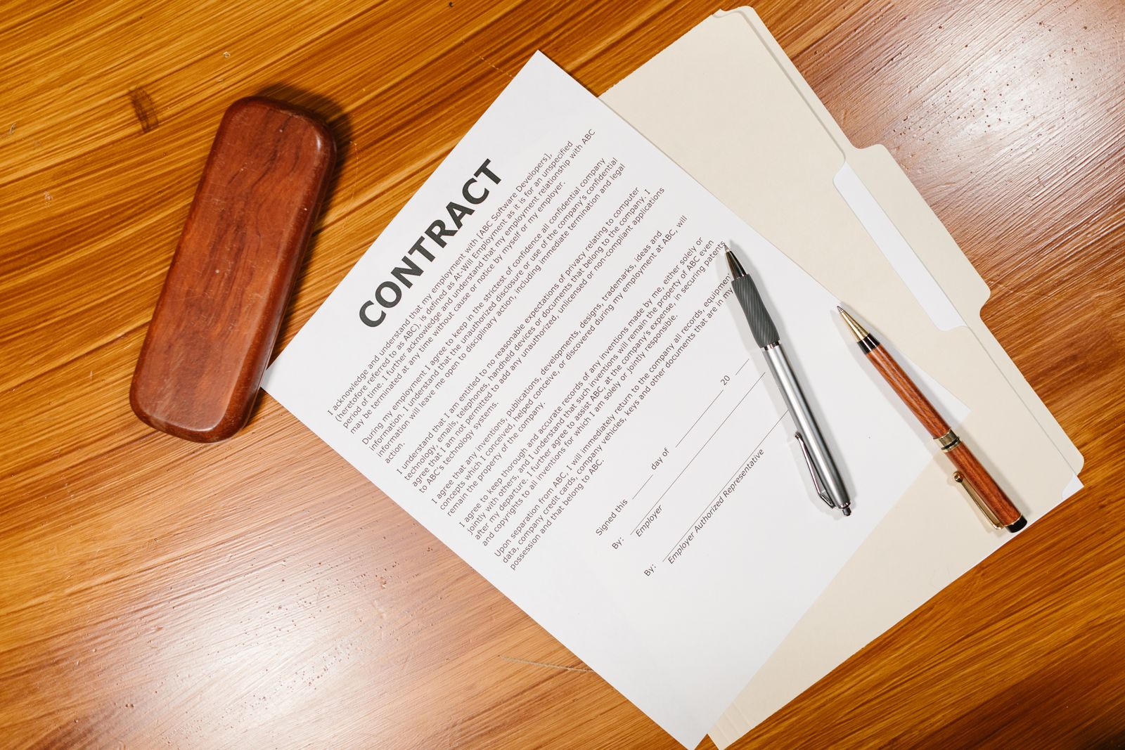 Printed contract pages spread on a wooden table with two pens and a document case