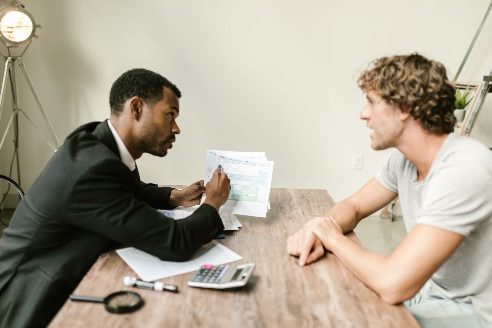 A man in a suit holding a printed document across a wooden table while another man listens, with a calculator and papers between them
