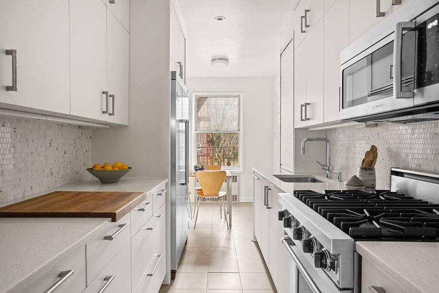 windowed eat-in kitchen filled with sleek white cabinetry and countertops, natural marble mosaic backsplash and upscale stainless steel appliances