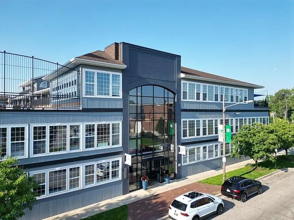 Elevated view of the three‑story Seventh Street Lofts condo building in Grand Rapids with gray‑blue siding, rows of white‑framed windows, a central glass entrance, rooftop railing, and cars parked along the street under a clear sky.