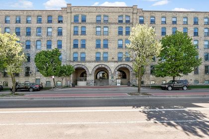 Wide frontal view of Boardwalk Condos, a long blonde‑brick warehouse‑turned‑condo building with arched stone entryways and rows of blue‑framed windows.