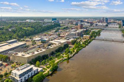 Drone view looking south over the Grand River with Boardwalk Condos occupying a full‑block brick complex at the river’s edge, downtown Grand Rapids skyline in the distance.