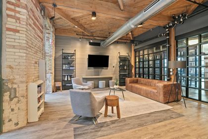 tylish community lounge with exposed brick walls, timber beams, leather sofa, gray armchairs, mounted flat‑screen TV, and industrial ductwork overhead.