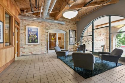 Ground‑floor lobby area with tiled floor, three gray armchairs around a glass coffee table, exposed brick walls, large arched window, and wood‑beam ceiling with ductwork.