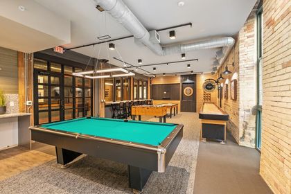Brick‑walled condo game room featuring a billiards table in the foreground, shuffleboard and foosball tables behind it, dartboards on the rear wall, exposed ductwork, and industrial lighting.