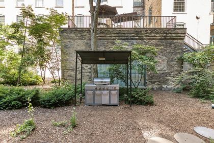 Covered stainless gas grill set beneath a metal canopy in a wooded condo courtyard, surrounded by mulch, shrubs, and historic brick walls.