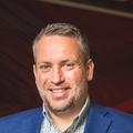 Close‑up headshot of a smiling man with short salt‑and‑pepper hair and trimmed beard, wearing a blue blazer and check‑pattern shirt against a dark red background.