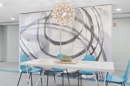 White rectangular table with four blue chairs beneath a latticed pendant light, backed by a sweeping black‑and‑gray abstract mural in the Seventh Street Lofts common area.