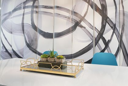 Close‑up of a mirrored gold tray holding small succulent planters on a glossy white table; abstract gray brushstroke mural and a blue chair appear softly in the background.