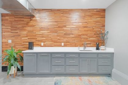 Communal kitchenette featuring blue‑gray lower cabinets with silver pulls, white quartz countertop, stainless sink, and a honey‑toned wood‑slat accent wall.