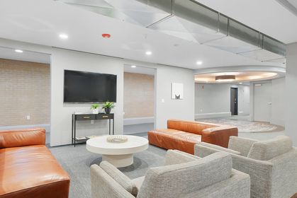 Common lounge area with tan leather couches, gray armchairs, a round white coffee table, wall‑mounted TV, exposed metal ducting on the ceiling, and hallway beyond.