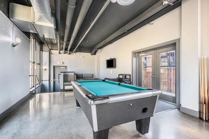 Industrial‑style game room with a pool table, TV, gray seating booths, and exposed ductwork ceiling.