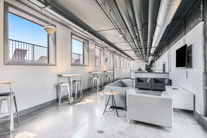 Long, light‑filled lounge with café tables, bar stools, gray sectional seating, TV, and exposed ceiling pipes.