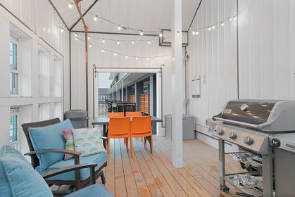 deck2Enclosed rooftop deck with a gas grill, wooden plank floor, blue cushioned wicker chairs, small dining table with two bright orange chairs, and string lights overhead.