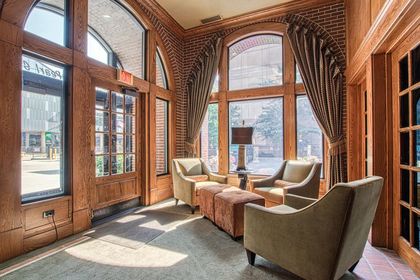Interior seating area with two armchairs, ottomans, tall arched windows, brick walls, and heavy drapery.