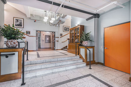 Lobby with terrazzo steps, vintage photos on blue walls, orange door, exposed pipes, and wood cabinets.