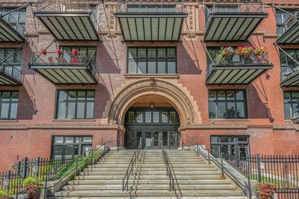 Front view of broad stone steps leading to a carved brick arch entry, framed by steel‑and‑glass balconies overhead.