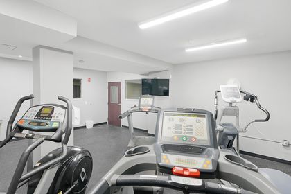 Modern apartment fitness room with gray walls featuring treadmills and elliptical machines in the foreground, wall‑mounted TV in the back, and rubber flooring.