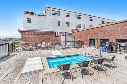 Small rectangular hot‑tub pool on a wooden rooftop deck, chairs alongside, brick and metal siding in the background.
