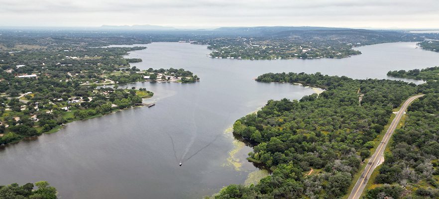 Aerial view of Inks Lake showing a boat leaving a wake in the lowered water, surrounded by green wooded areas and residential properties, with a road visible alongside the shoreline.